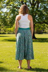Person wearing a white tank top and green patterned skirt in a sunny park with a big tree