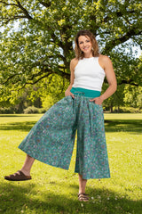Woman wearing teal floral wide-leg pants and a white top ina a park with a big tree