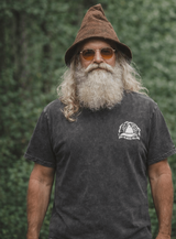 Man wearing a black t-shirt with a gnome graphic and text, standing against a green forest background.