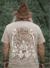 Man wearing a beige t-shirt with a gnome graphic and text, standing against a green leafy background.
