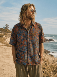 A man with long blonde hair and sunglasses stands on a sandy beach wearing a short-sleeve button-up shirt with an intricate multicolor pattern in blue, orange, and brown, paired with olive green pants.