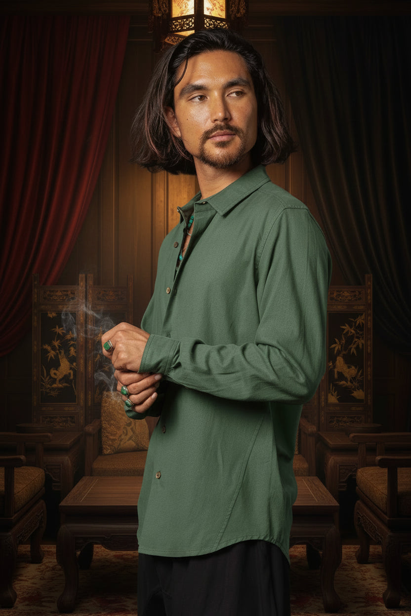 A man with long dark hair and a beard wearing an olive green long-sleeve button-up shirt, standing against a dark textured background.