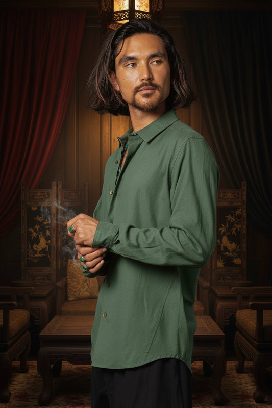 A man with long dark hair and a beard wearing an olive green long-sleeve button-up shirt, standing against a dark textured background.