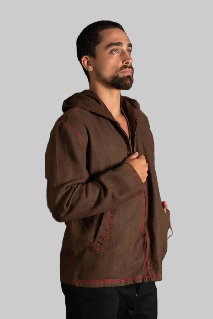 A man wearing a brown hooded jacket with red contrast stitching, standing against a dark textured background.