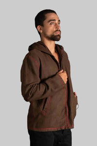 A man wearing a brown hooded jacket with red contrast stitching, standing against a dark textured background.