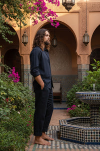 A man with long brown hair and a beard stands barefoot against a dark textured backdrop, wearing a loose black long-sleeve button-up shirt and matching black pants.