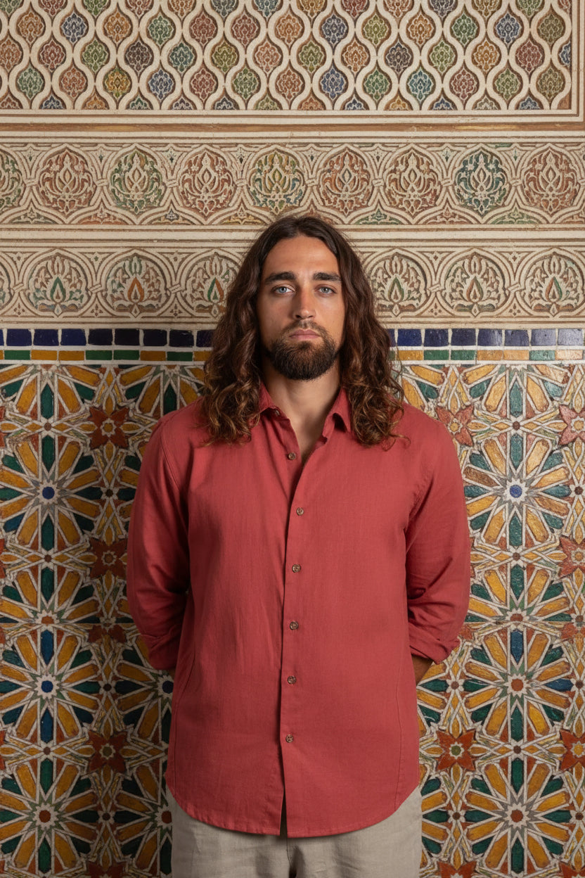 A man with long brown hair and a beard wearing a muted red button-down shirt and beige pants, standing against a dark textured background.