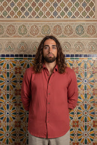 A man with long brown hair and a beard wearing a muted red button-down shirt and beige pants, standing against a dark textured background.