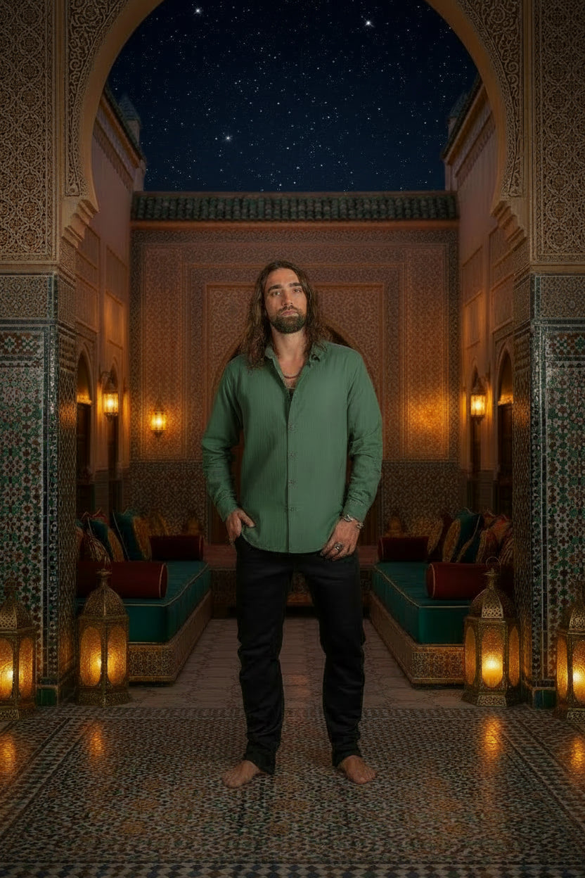 A man with long brown hair and a beard wearing a muted sage green long-sleeve button-up shirt and black pants, standing barefoot against a dark textured studio backdrop.