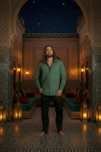 A man with long brown hair and a beard wearing a muted sage green long-sleeve button-up shirt and black pants, standing barefoot against a dark textured studio backdrop.