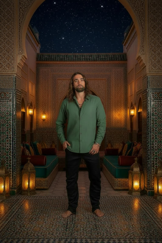A man with long brown hair and a beard wearing a muted sage green long-sleeve button-up shirt and black pants, standing barefoot against a dark textured studio backdrop.