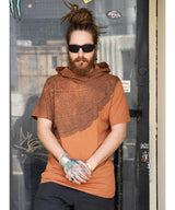 Man wearing a brown patterned hoodie over an orange t-shirt in front of a glass door.