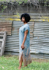 Woman in a long, light blue dress standing in front of a corrugated metal wall.