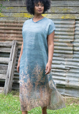 Woman wearing a blue patterned dress standing in front of a rustic wooden wall.