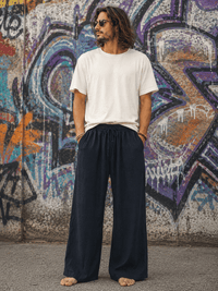 A man with long dark hair and sunglasses stands barefoot in front of a graffiti-covered wall, wearing a plain white short-sleeve t-shirt and dark navy wide-leg drawstring pants.