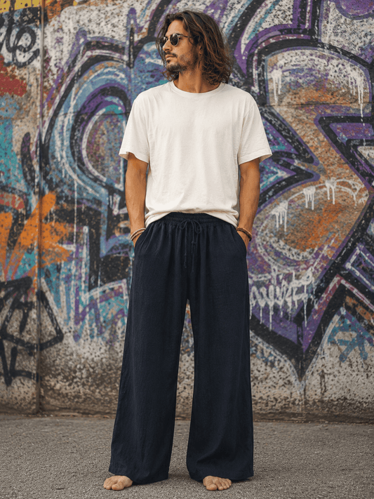 A man with long dark hair and sunglasses stands barefoot in front of a graffiti-covered wall, wearing a plain white short-sleeve t-shirt and dark navy wide-leg drawstring pants.