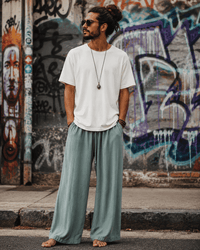 A man wearing a white t-shirt, sage green wide-leg pants, and sunglasses stands barefoot on a street in front of a graffiti-covered wall.