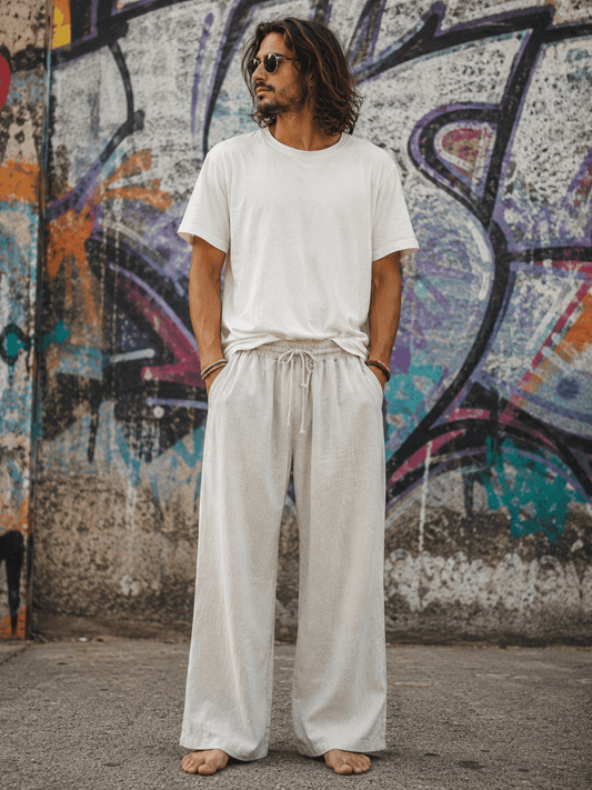 A man wearing a white crew-neck t-shirt and matching white wide-leg linen pants, standing barefoot in front of a colorful graffiti wall.