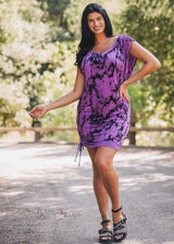 Woman wearing a purple tie-dye dress standing outdoors with trees in the background