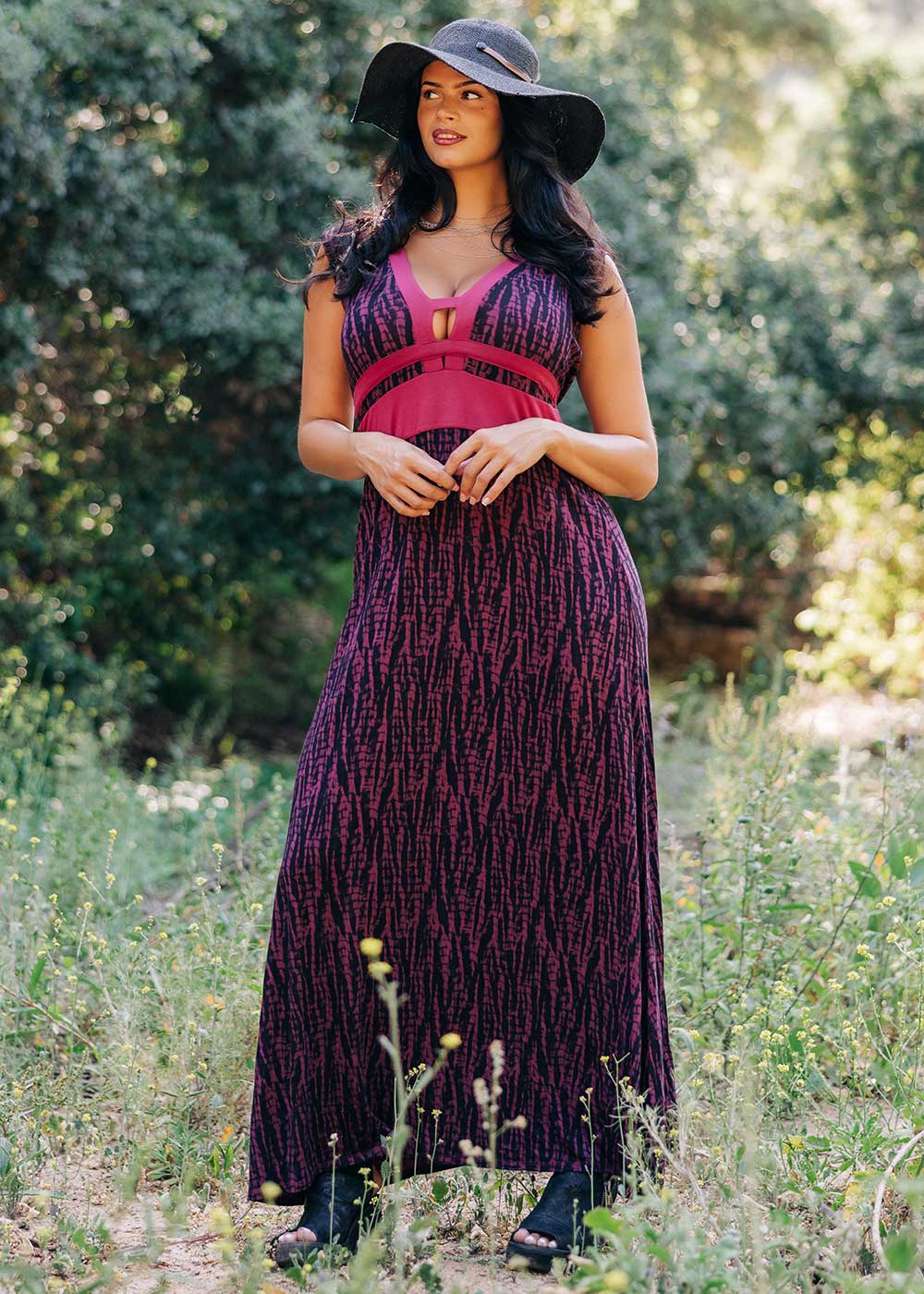 Woman in a long, dark patterned dress and wide-brimmed hat standing in a natural setting.
