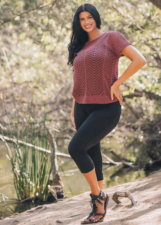 Woman wearing a maroon knitted top and black leggings standing outdoors by a body of water.