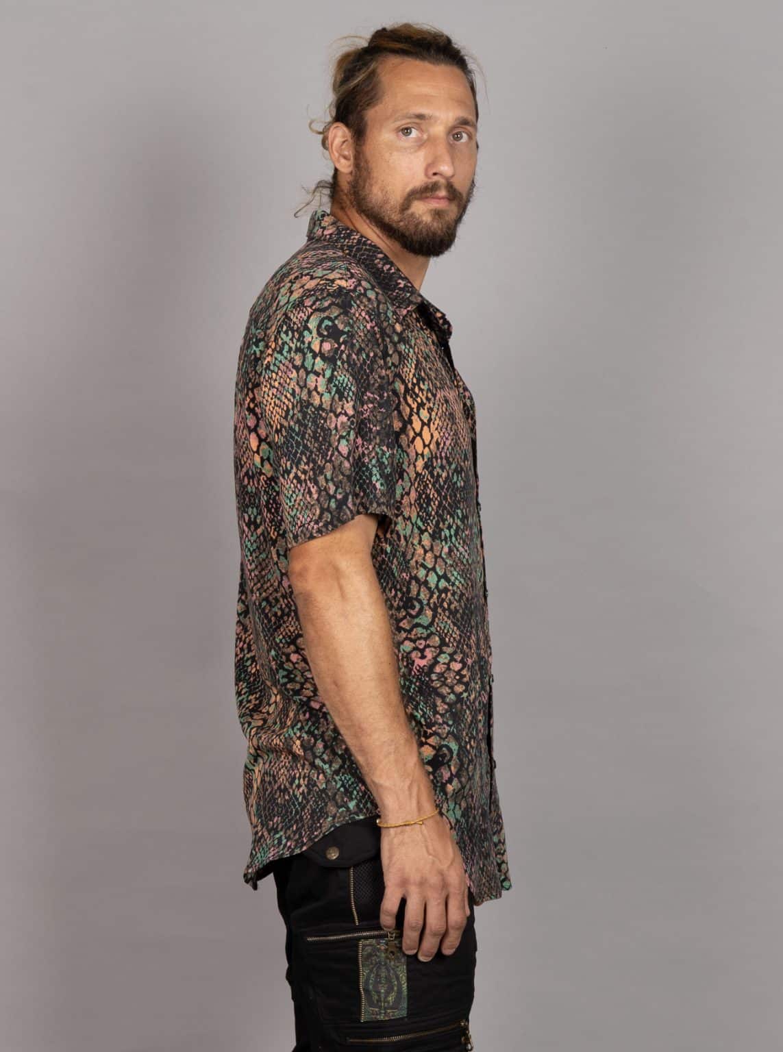 A man wearing a short-sleeve button-up shirt with a complex multicolor snakeskin pattern, paired with black jeans, standing against a solid gray studio background.