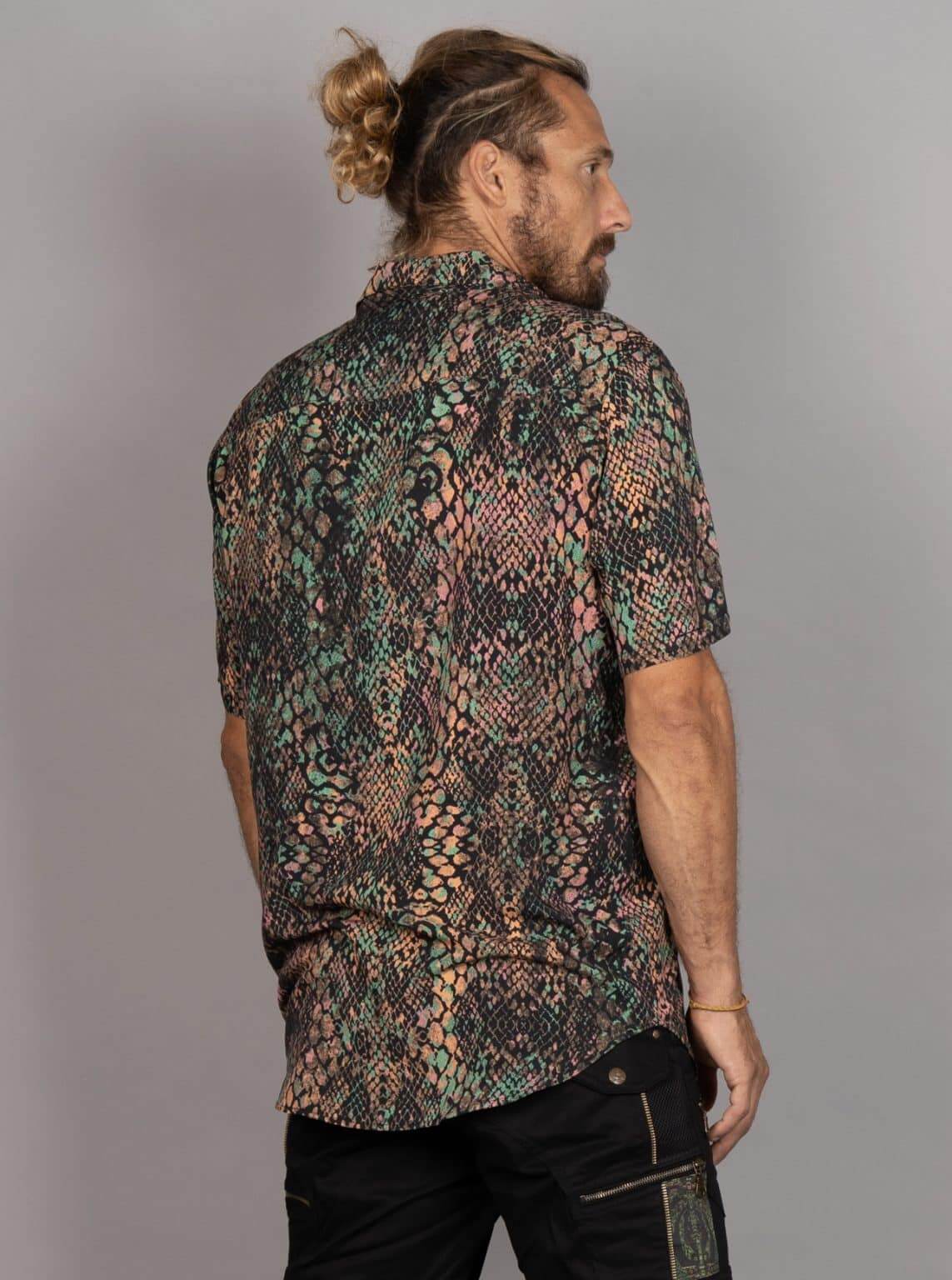 A man with a bun and beard, shown from the back, wearing a short-sleeve button-up shirt with a complex multicolor snakeskin pattern in green, pink, black, and beige, paired with black pants, against a plain gray background.