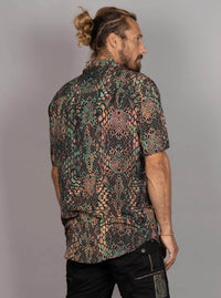 A man with a bun and beard, shown from the back, wearing a short-sleeve button-up shirt with a complex multicolor snakeskin pattern in green, pink, black, and beige, paired with black pants, against a plain gray background.