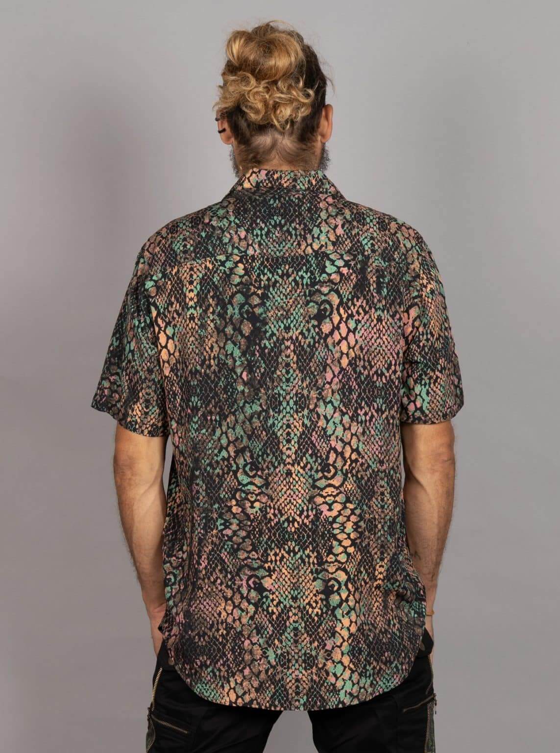 A man viewed from behind wearing a short-sleeve button-up shirt with a complex multicolor snakeskin pattern in black, green, and peach tones, paired with black pants against a solid gray background.