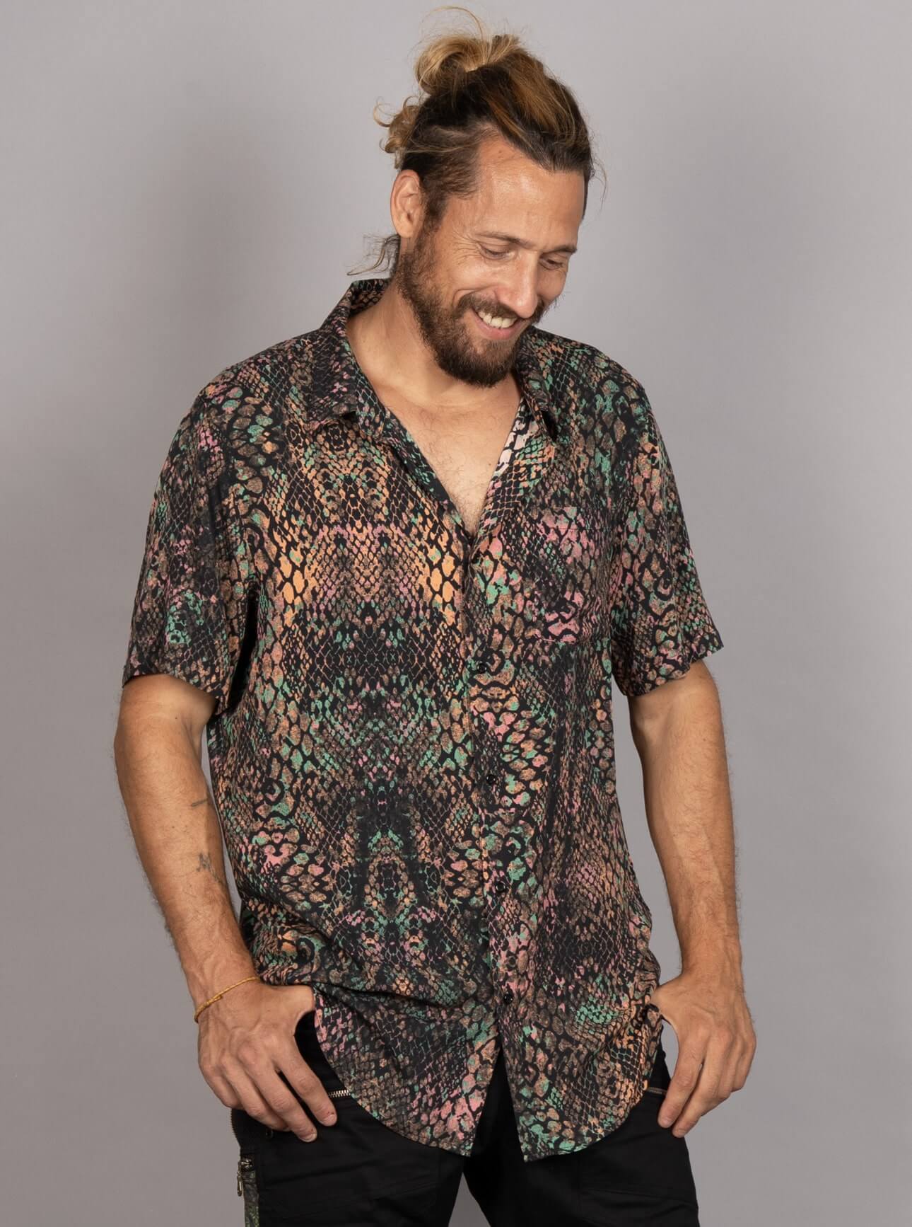 A man wearing a short-sleeve button-up shirt with a multicolor snakeskin pattern in black, green, pink, and beige, paired with black pants, set against a plain gray studio background.