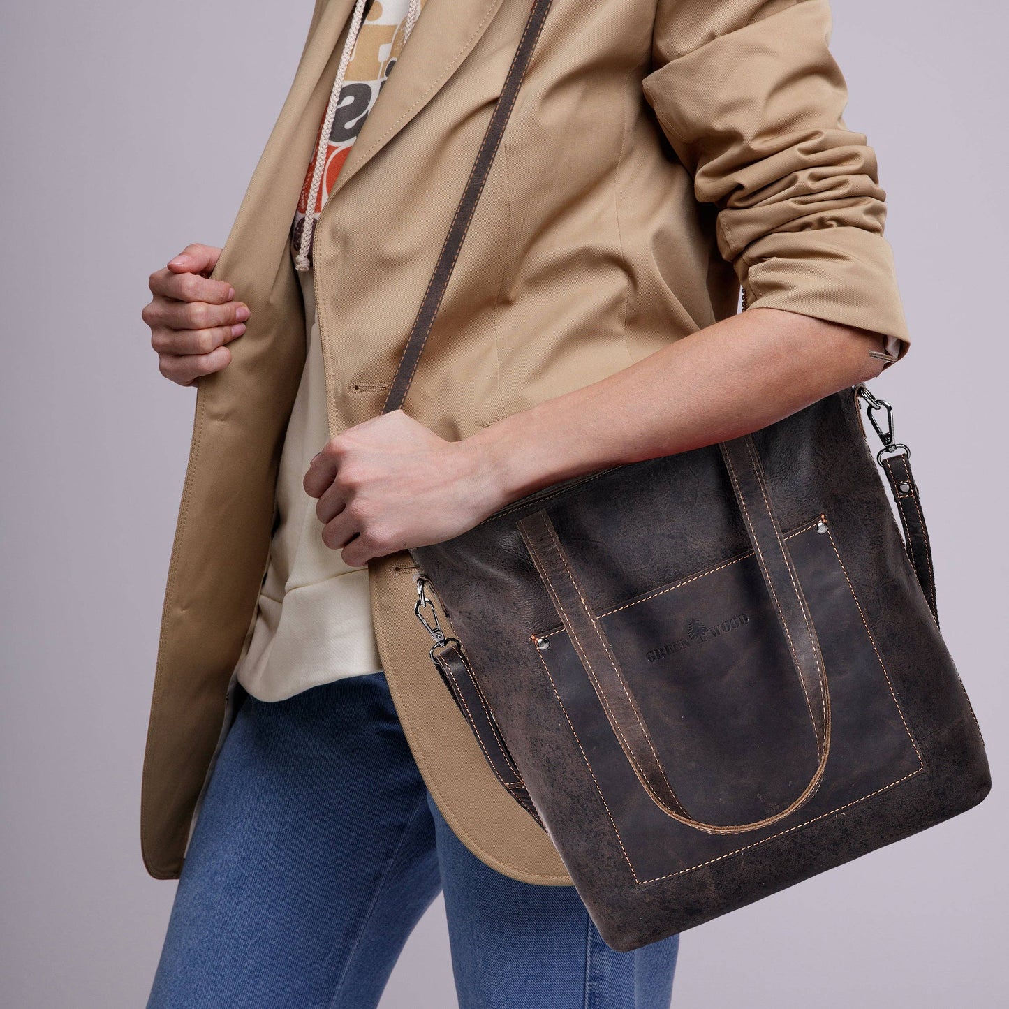 Person wearing a beige jacket and blue jeans holding a dark brown leather bag against a plain background