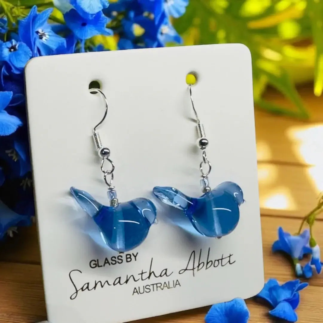 Blue glass bird earrings on a branded card with flowers in the background