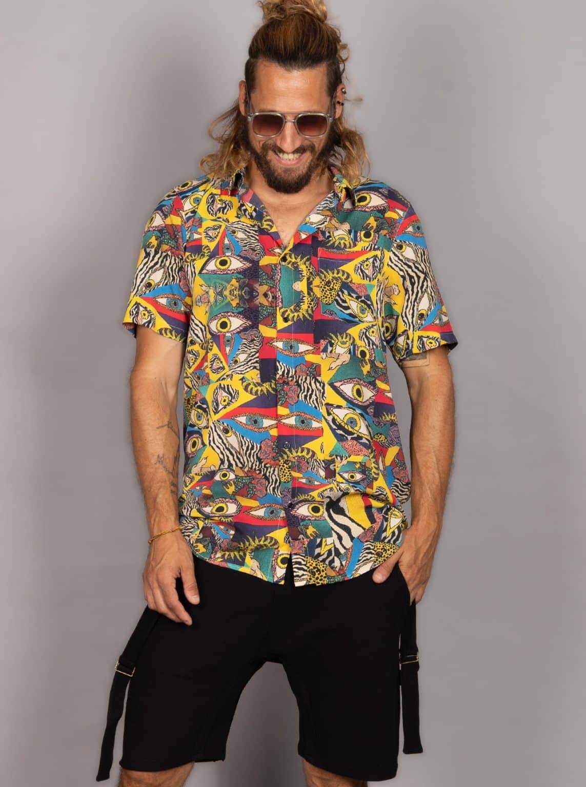 A man wearing a short-sleeve button-up shirt with a vibrant multicolor abstract print featuring eyes, stripes, and geometric shapes, paired with black cargo shorts, set against a plain gray studio background.