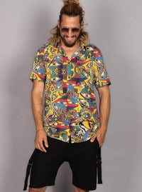 A man wearing a short-sleeve button-up shirt with a vibrant multicolor abstract print featuring eyes, stripes, and geometric shapes, paired with black cargo shorts, set against a plain gray studio background.