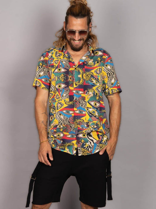 A man wearing a short-sleeve button-up shirt with a vibrant multicolor abstract print featuring eyes, stripes, and geometric shapes, paired with black cargo shorts, set against a plain gray studio background.