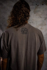Person wearing a brown t-shirt with a logo on the back against a textured wall.