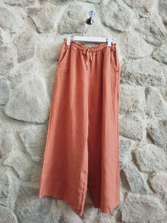 A pair of wide-leg linen pants in a warm terracotta or rust orange color, hanging from a white plastic hanger against a textured stone wall.