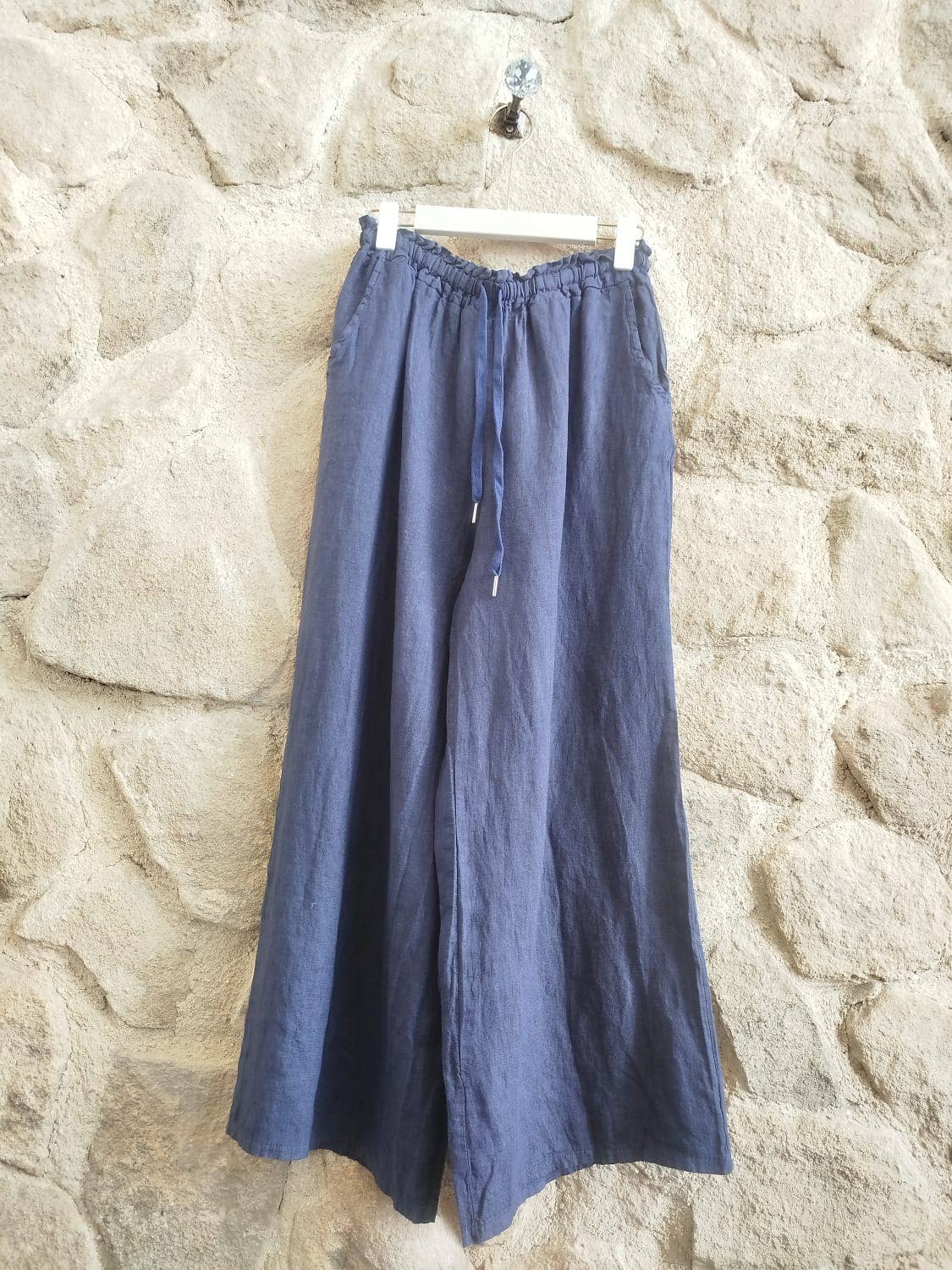 A pair of wide-leg linen pants in a muted slate blue color, hanging from a white plastic hanger against a rustic stone wall.