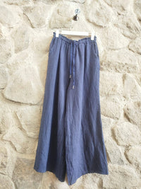 A pair of wide-leg linen pants in a muted slate blue color, hanging from a white plastic hanger against a rustic stone wall.