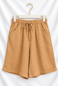 A pair of tan linen shorts with an elastic drawstring waist and side pockets, hanging on a white hanger with a gold hook against a white fabric backdrop.