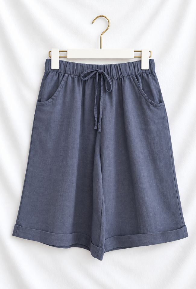A pair of slate blue linen shorts with an elastic drawstring waist and side pockets, hanging on a white hanger against a white fabric backdrop.