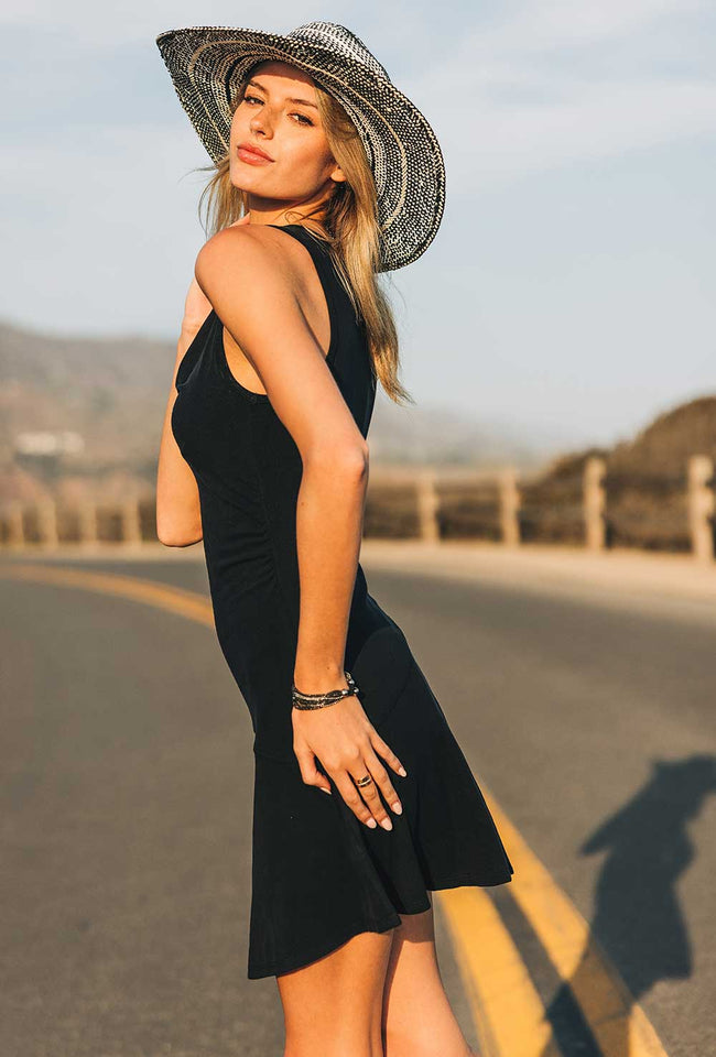 Woman in a black dress and straw hat standing on a road with mountains in the background