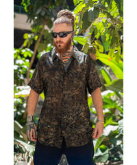 Man wearing a patterned shirt and sunglasses standing in front of green foliage
