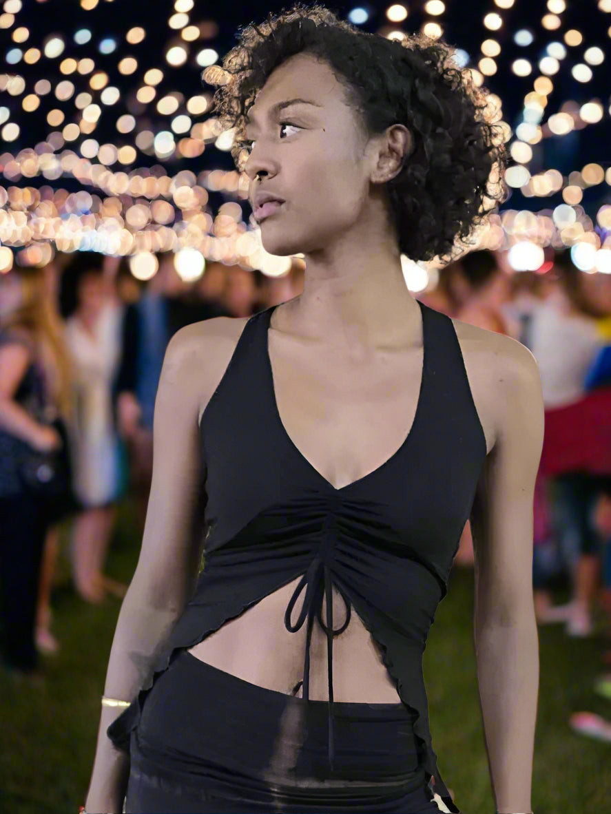 A woman wearing a black tie-up dance top with adjustable straps and a front tie, standing in a park with festive lights in the background.