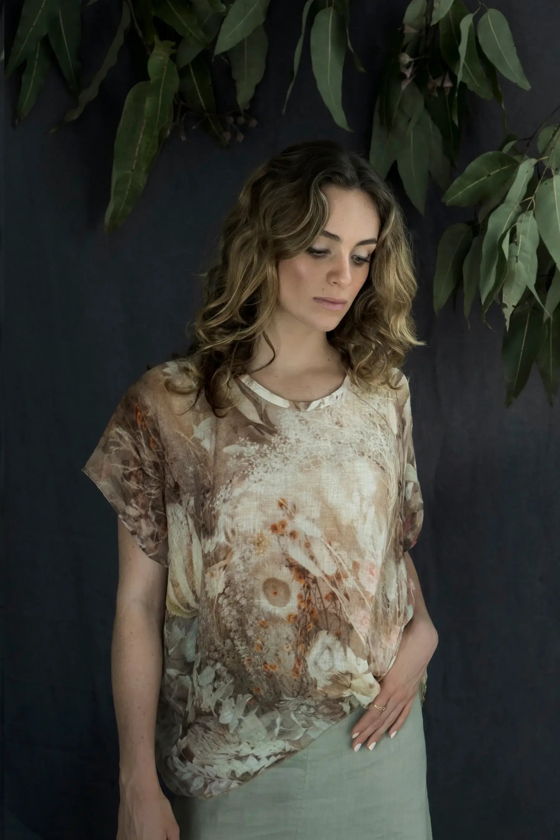 Woman wearing a patterned shirt against a dark background with greenery.