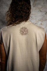 Person wearing a beige sleeveless shirt with a logo on the back against a textured wall.