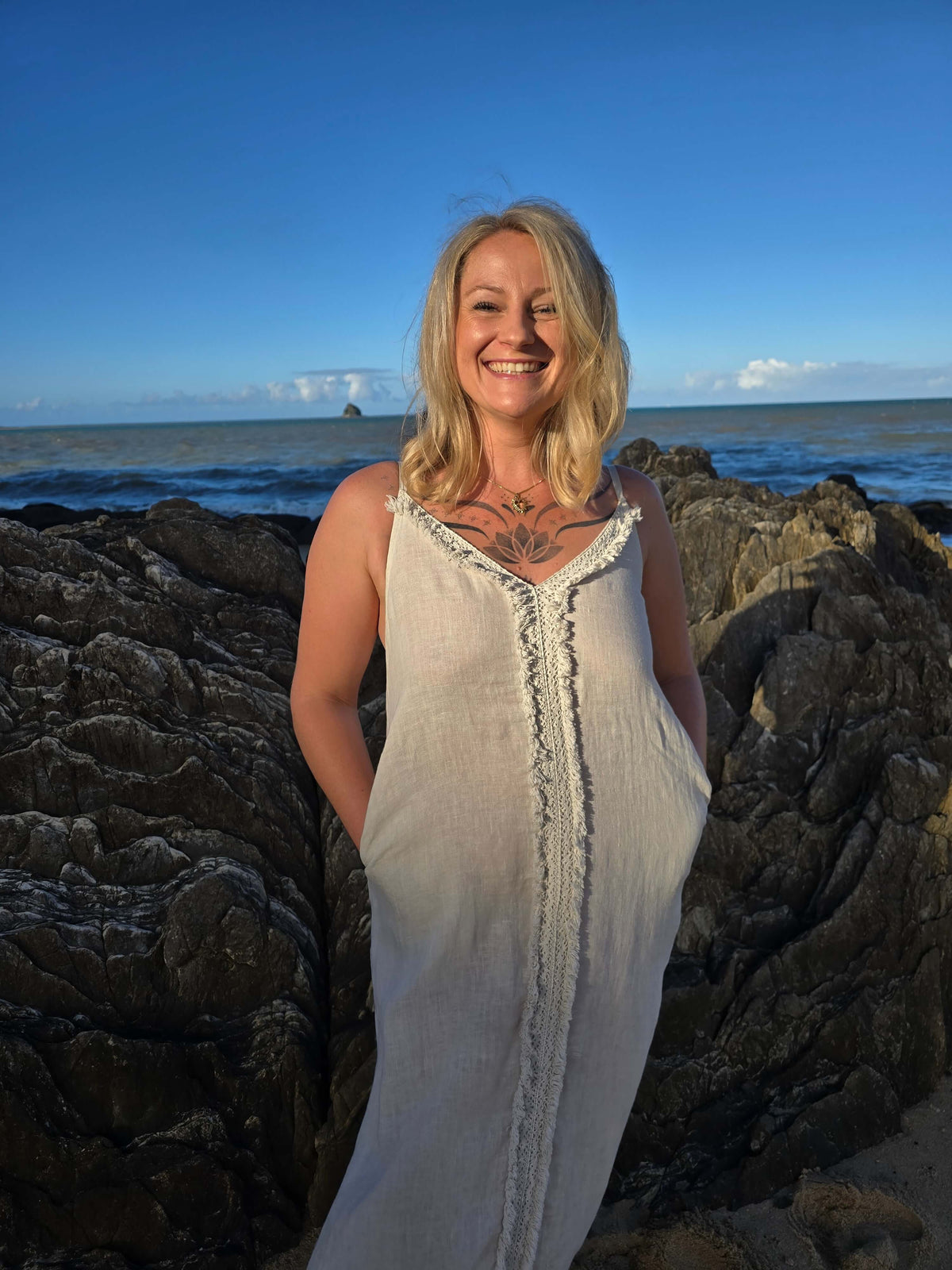 A smiling blonde woman wearing a sleeveless cream linen dress stands on dark rocks with the ocean and blue sky in the background.