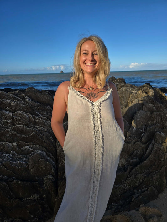 A smiling blonde woman wearing a sleeveless cream linen dress stands on dark rocks with the ocean and blue sky in the background.