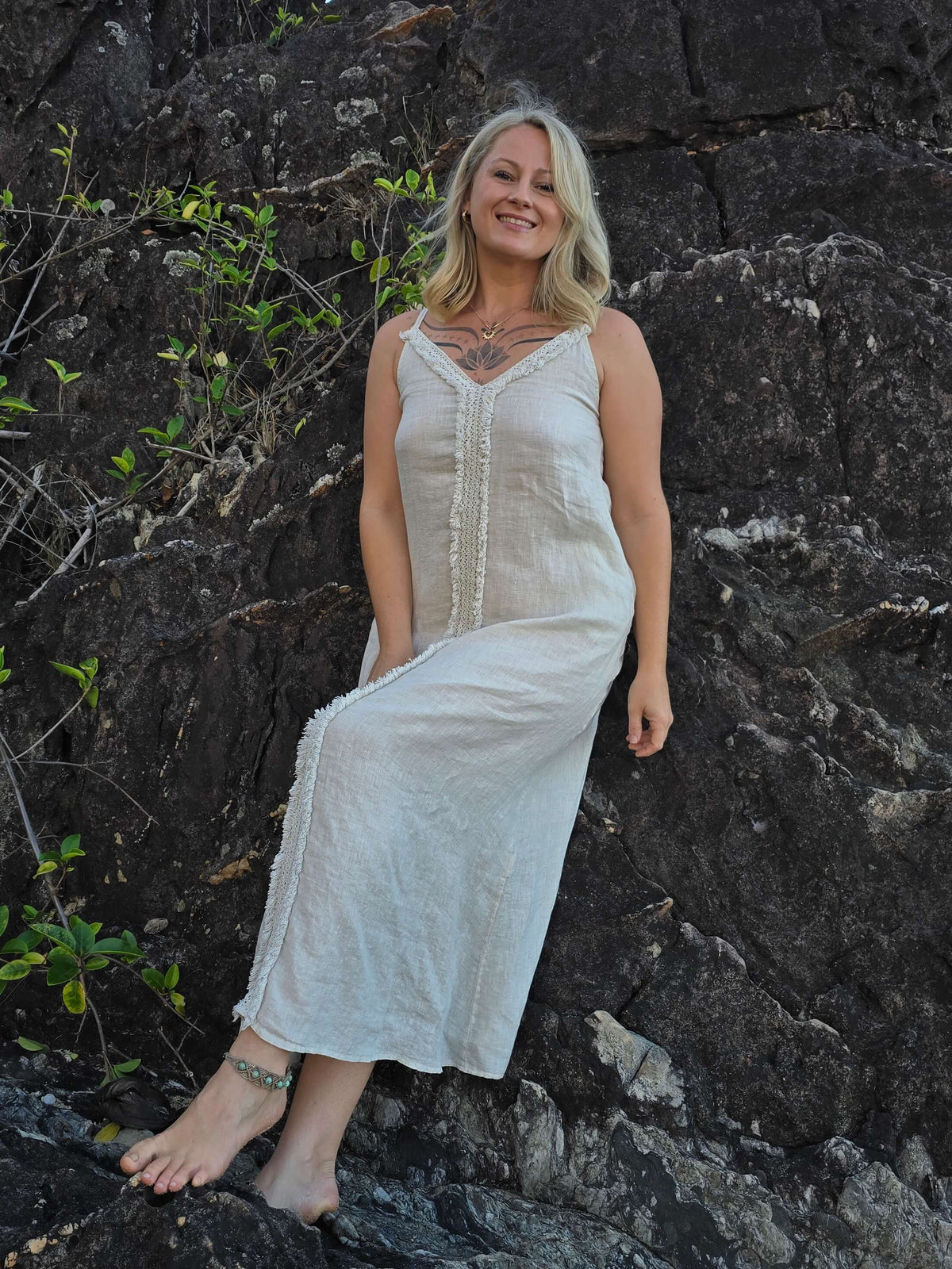 A woman wearing a sleeveless, off-white maxi dress with a lace-up front, posing on dark volcanic rocks outdoors.