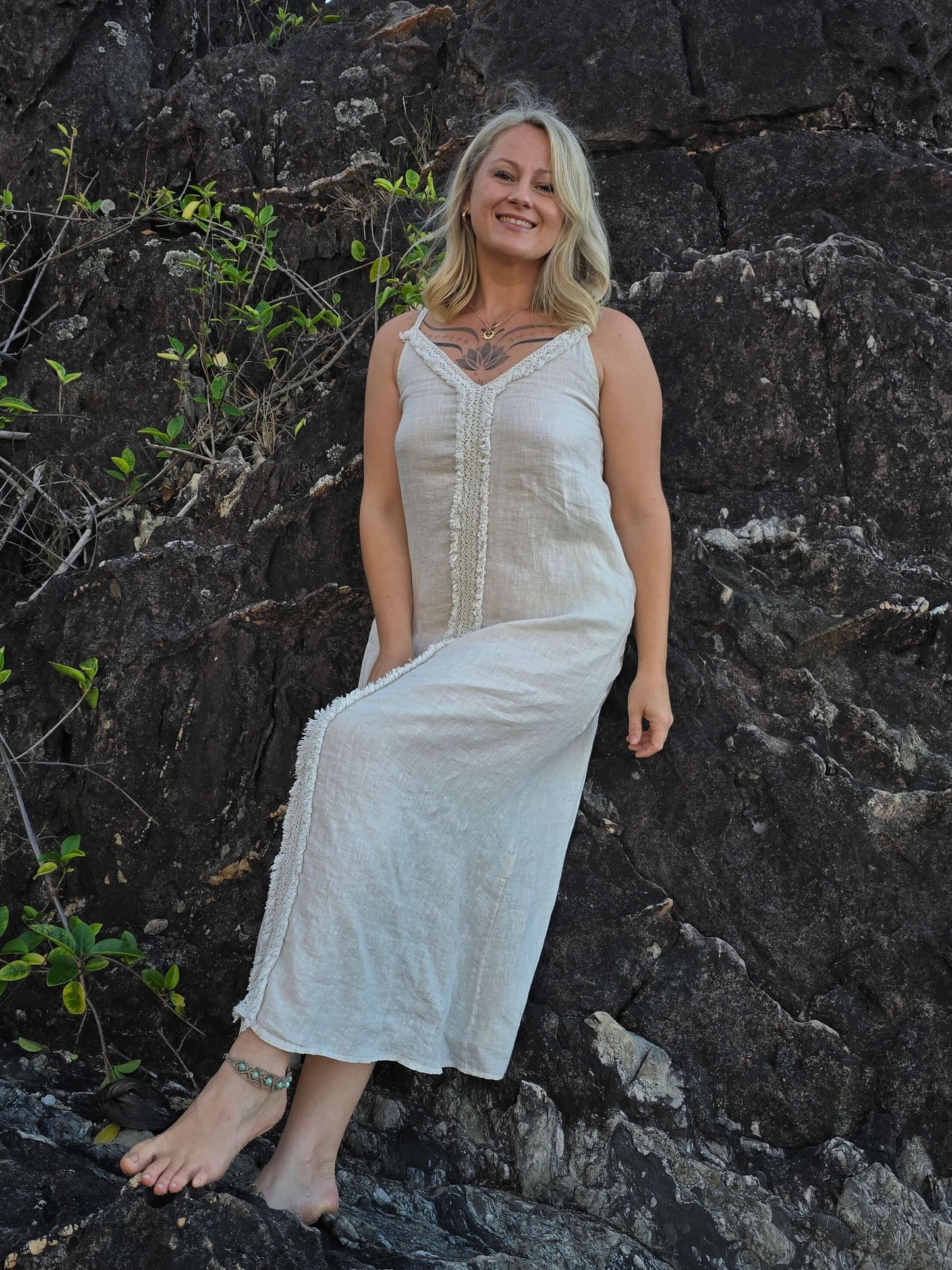 A woman wearing a sleeveless, off-white maxi dress with a lace-up front, posing on dark volcanic rocks outdoors.